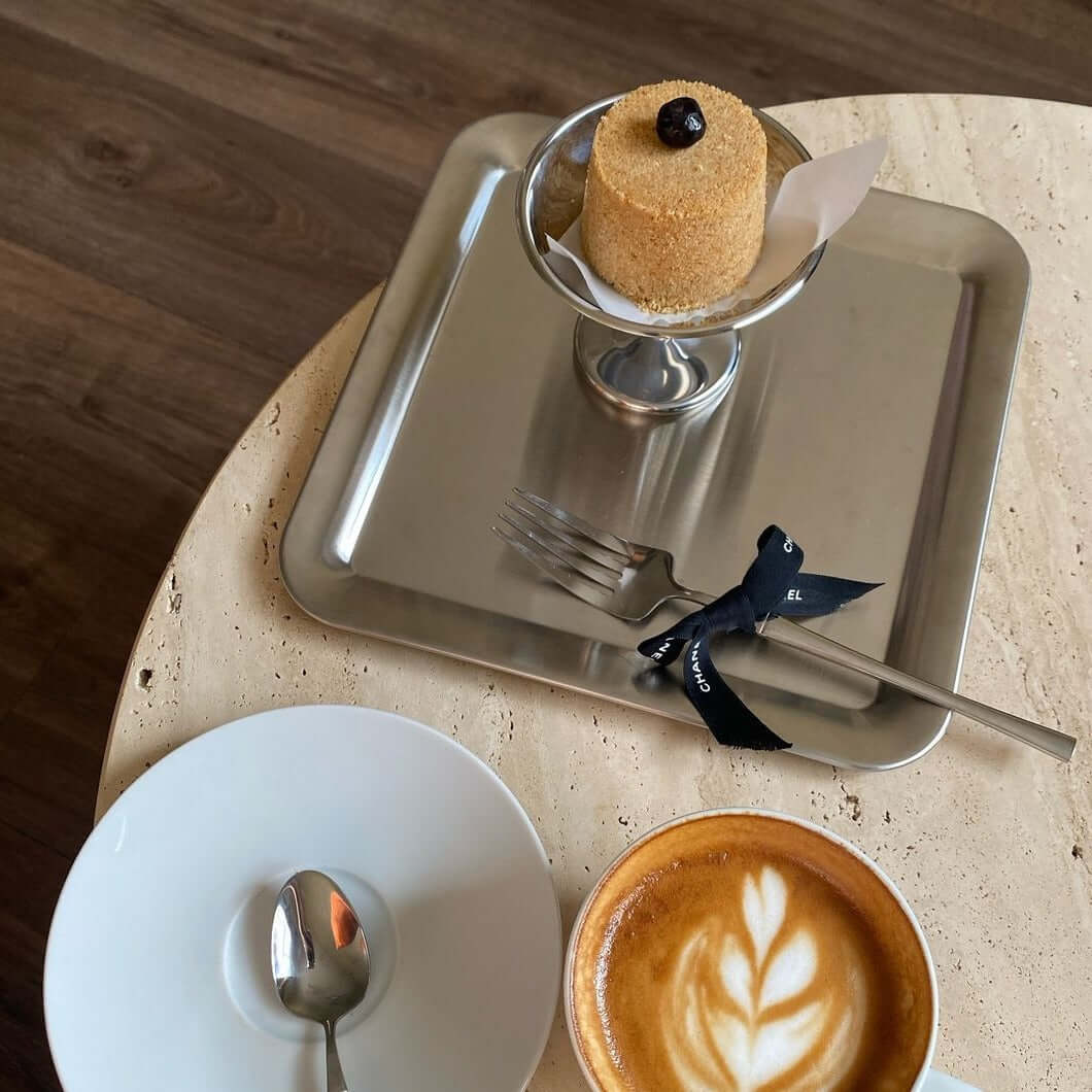 Urban Chic stainless steel serving tray with elegant dessert display, alongside a cup of cappuccino and a white saucer on a table.