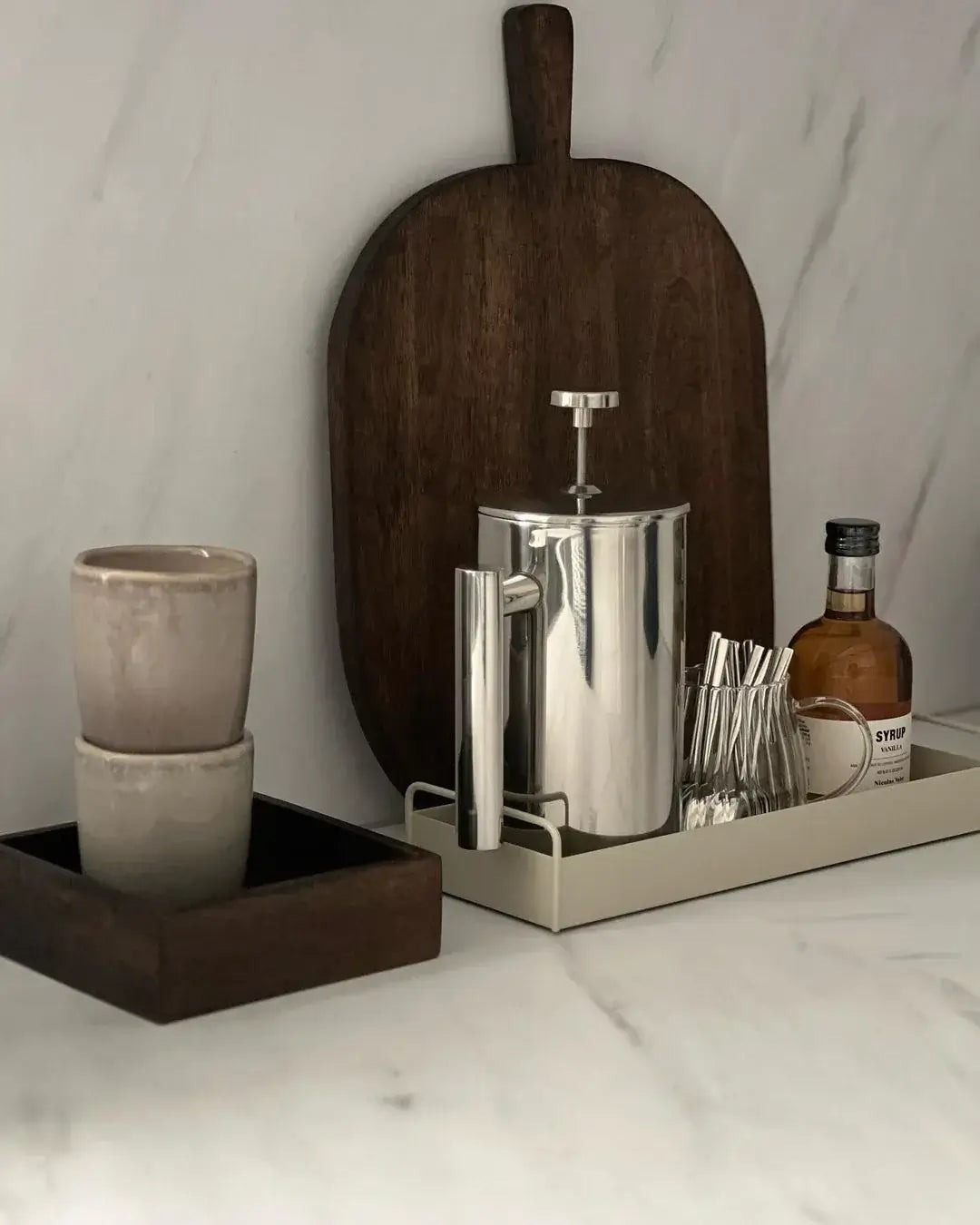 Sleek stainless steel French press with two cups and a wooden serving board on a marble countertop.