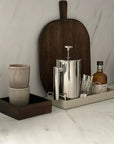 Sleek stainless steel French press with two cups and a wooden serving board on a marble countertop.