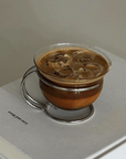 Muse Glass Cup with iced coffee on its scalloped steel saucer, resting on a book