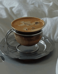 Muse Glass Cup & Saucer Set with an iced latte on its scalloped saucer, on a white bed