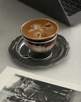 Muse Glass Cup & Saucer with iced coffee beside a black and white photo book