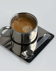 A stainless steel coffee cup with a mirror-like finish, accompanied by a matching saucer and spoon, containing a hot beverage.