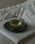 Muse Glass Cup with a clear drink on its scalloped saucer, beside a laptop