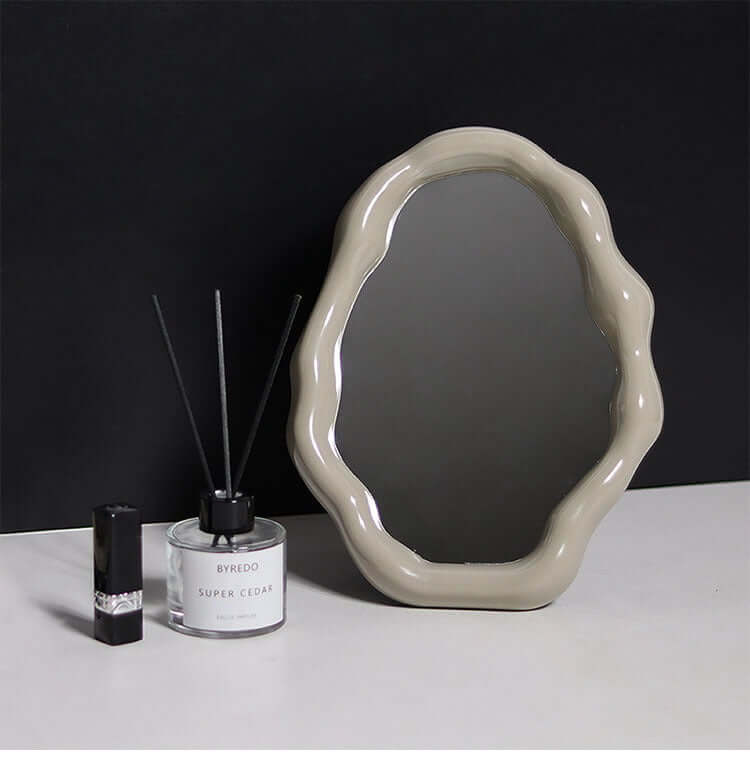 Cloud-shaped vanity mirror with a wavy beige frame on a black background, alongside a candle and perfume bottle.