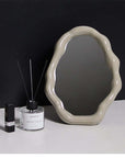 Cloud-shaped vanity mirror with a wavy beige frame on a black background, alongside a candle and perfume bottle.