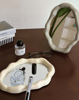 Nordic cloud-shaped vanity mirror on a wooden table with decor items including a book, glasses, and perfume bottle.