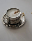 Stainless steel coffee cup with frothy coffee on saucer, part of Limited Steel Bundle set.