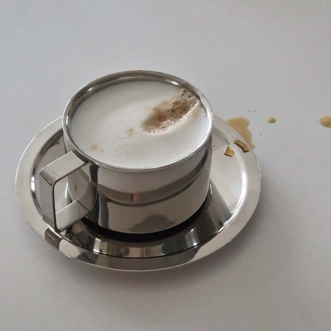 Italian style double-layered stainless steel latte cup with saucer, frothy coffee, and small spill. 200 ml capacity.