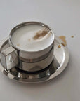Italian style double-layered stainless steel latte cup with saucer, frothy coffee, and small spill. 200 ml capacity.