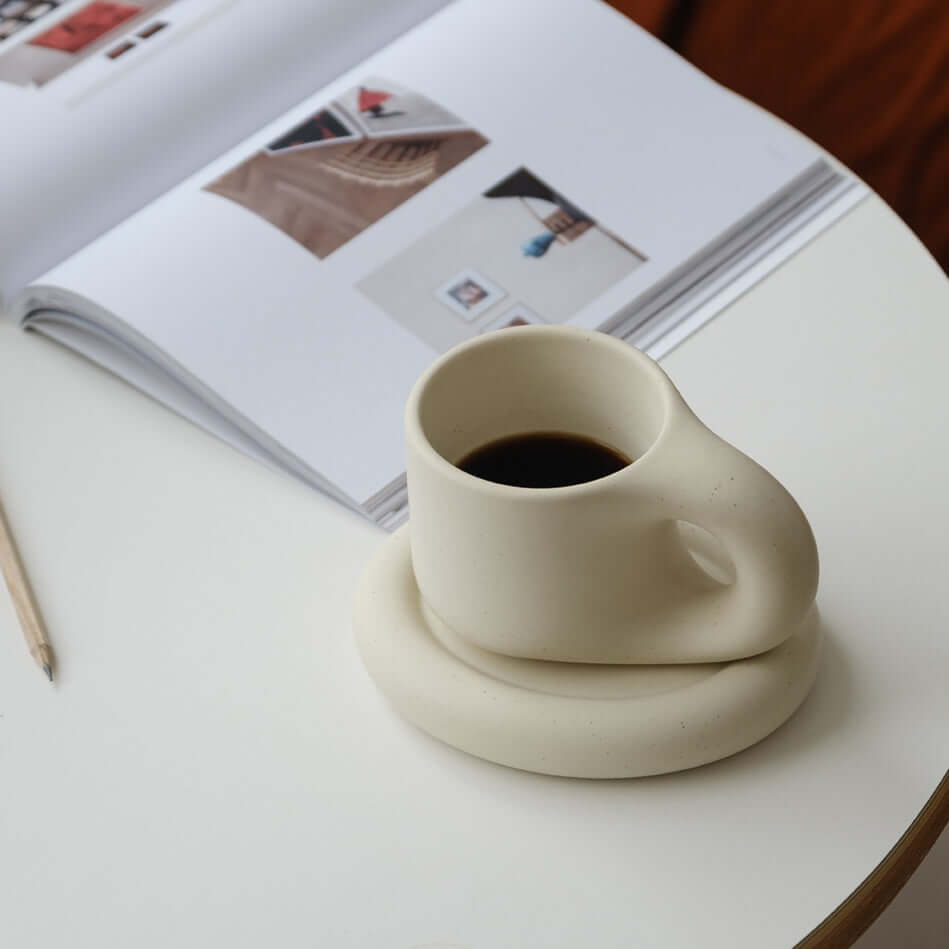 Artisan Nordic Chubby Mug & Plate with coffee on a table next to an open magazine, showcasing minimalist clay design.
