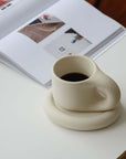 Artisan Nordic Chubby Mug & Plate with coffee on a table next to an open magazine, showcasing minimalist clay design.