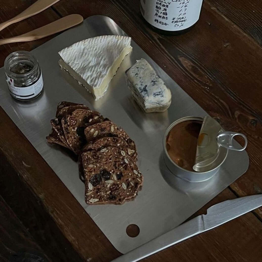 Minimalist stainless steel cutting board used as serving platter with cheese, crackers, and spread on wooden table.