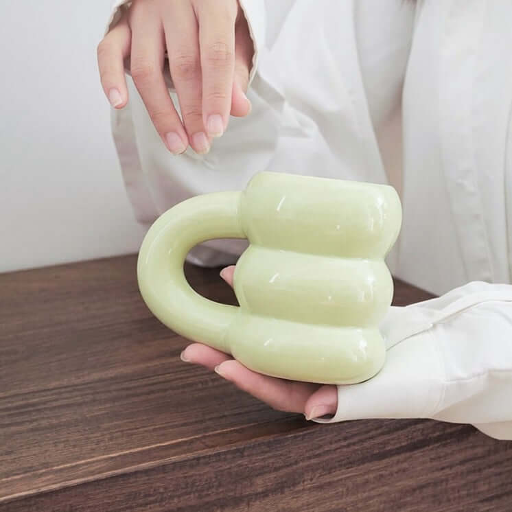 Unique green ceramic mug with a sculpted, modern design, held by a person on a wooden table.