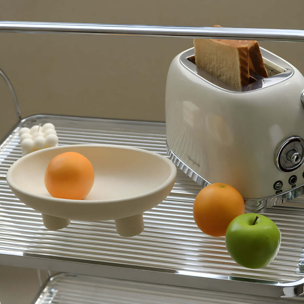 Ceramic three-legged fruit plate with oranges and a green apple beside a toaster on a shelf, enhancing modern kitchen decor.