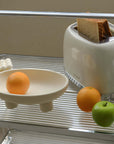 Ceramic three-legged fruit plate with oranges and a green apple beside a toaster on a shelf, enhancing modern kitchen decor.