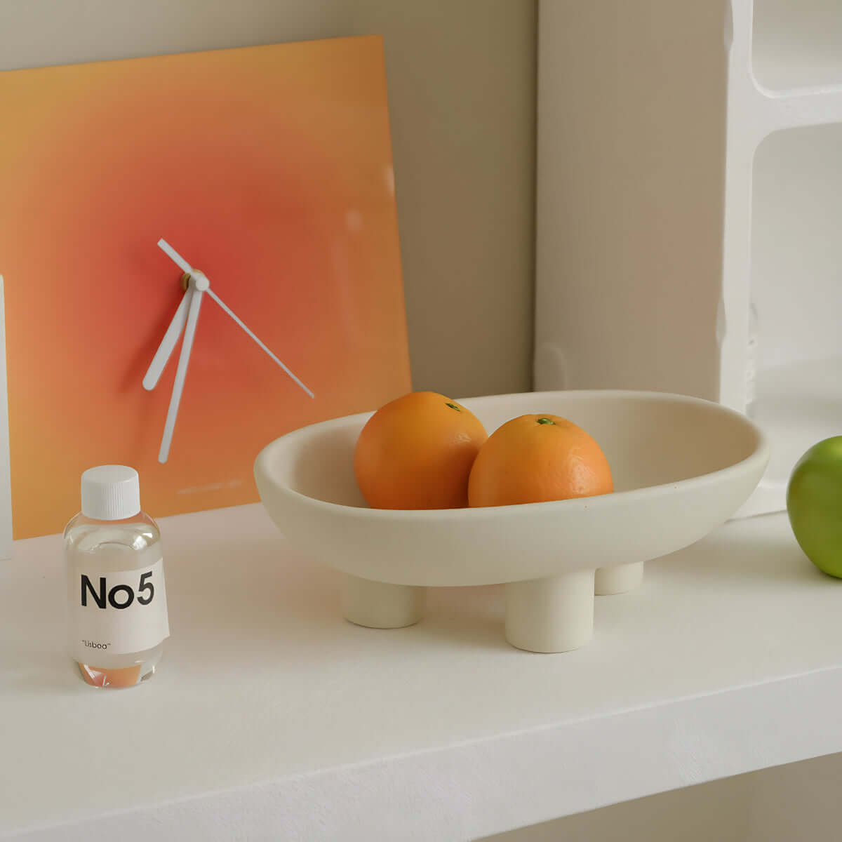 Ceramic three-legged plate with oranges, placed beside a modern clock and No5 bottle on a minimalist shelf.