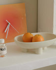 Ceramic three-legged plate with oranges, placed beside a modern clock and No5 bottle on a minimalist shelf.