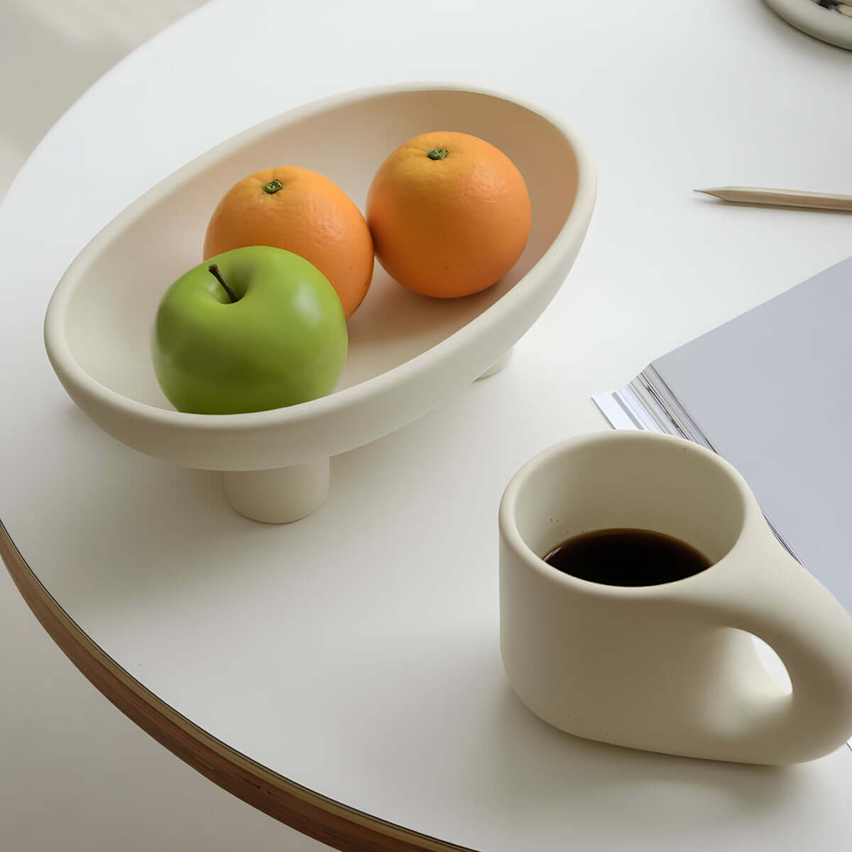 Ceramic three-legged fruit plate with oranges and an apple, next to a cup of coffee on a white table, enhancing modern dining decor.