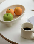 Ceramic three-legged fruit plate with oranges and an apple, next to a cup of coffee on a white table, enhancing modern dining decor.