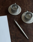 Stainless steel vintage cup and saucer set with pen on wooden table, showcasing elegant design and durability.