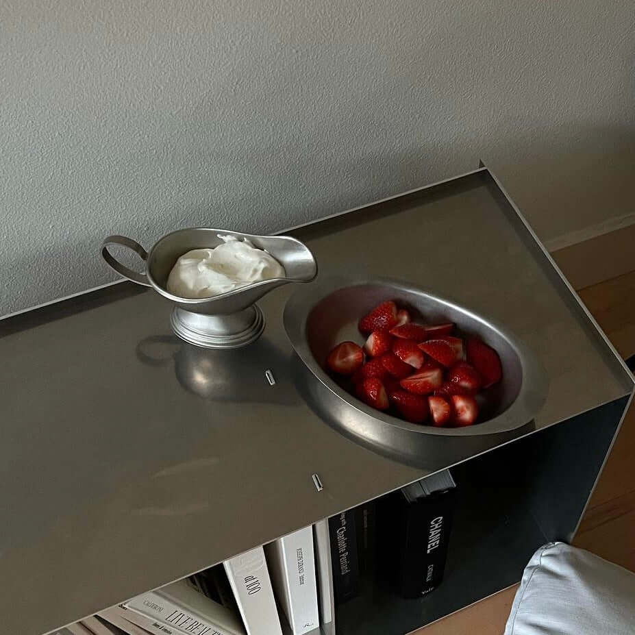 Stainless steel sauce pot with cream and bowl of strawberries on a metal surface.