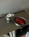 Stainless steel sauce pot with cream and bowl of strawberries on a metal surface.