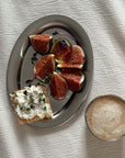Stainless steel plate with figs and cream cheese toast, accompanied by a cappuccino on a textured white fabric background.