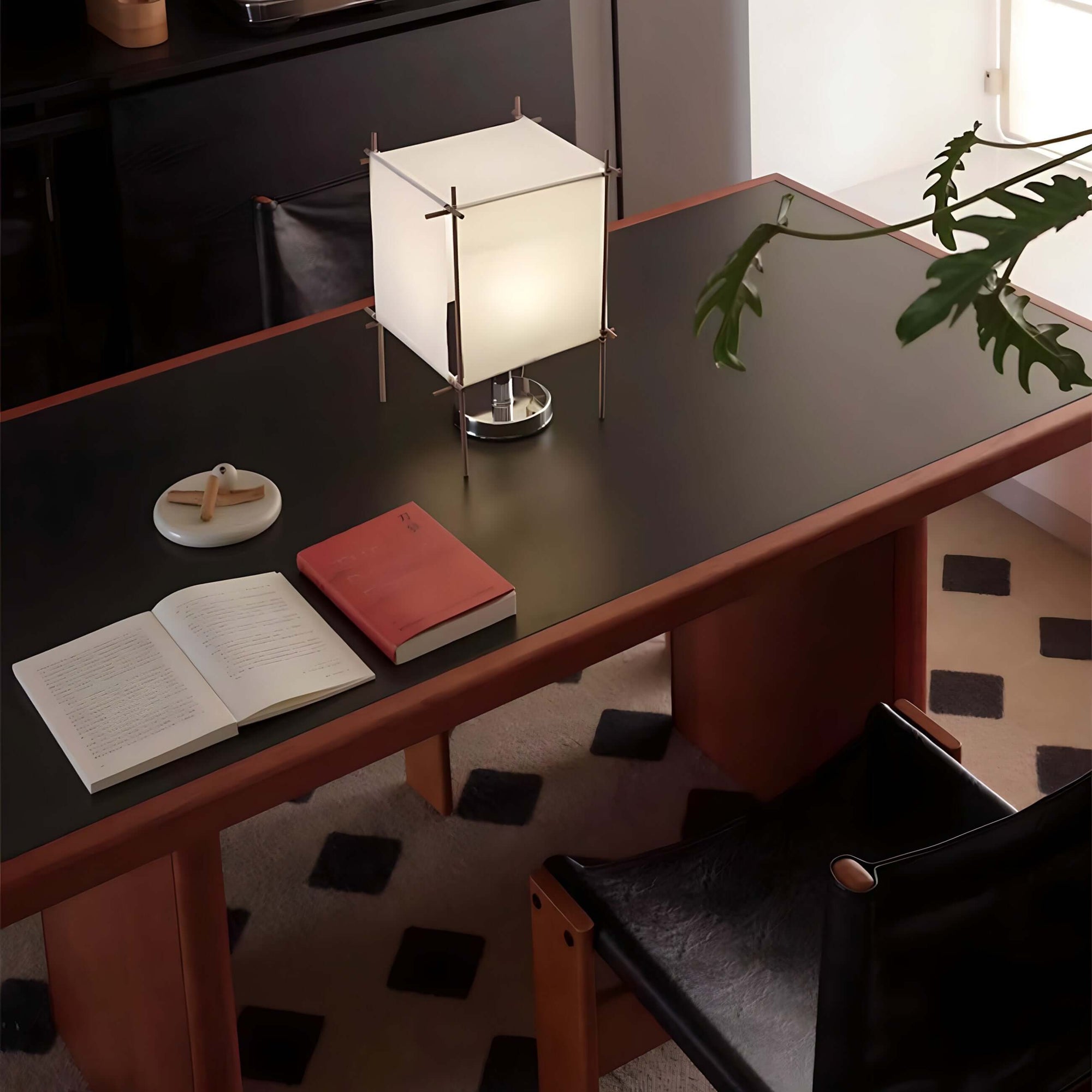 Elegant dining table setup with modern lamp, open book, and plant decor on a sleek dark surface.