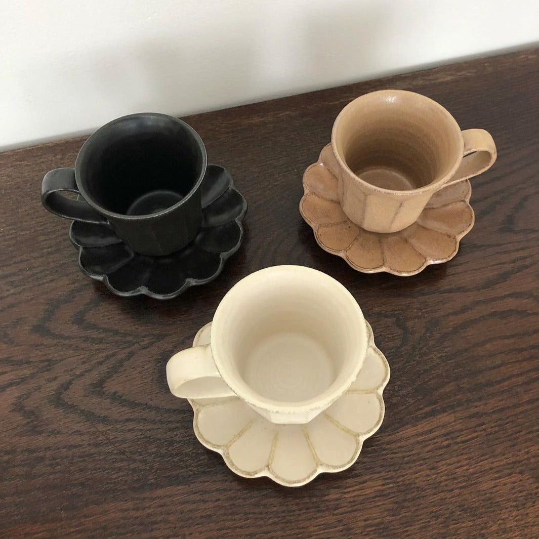Three Rinka porcelain mugs with flower-shaped saucers in black, brown, and ivory on a wooden table.