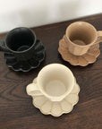 Three Rinka porcelain mugs with flower-shaped saucers in black, brown, and ivory on a wooden table.