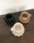 Rinka mugs with flower-shaped saucers in black, brown, and cream, handcrafted porcelain by Kaneko Kohyo on a wooden table.