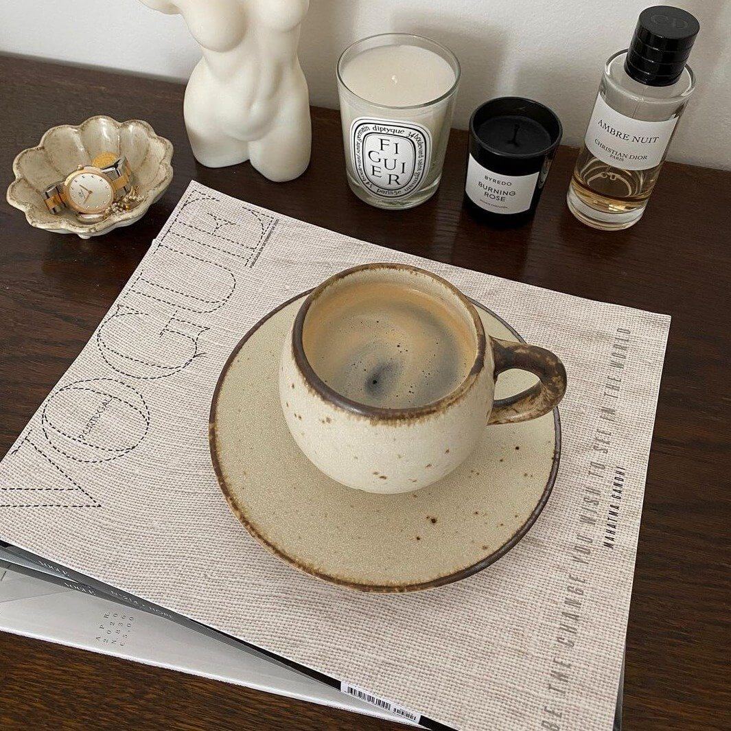 Rustic ivory mug on a saucer, placed on a fashion magazine, alongside candles and decor on a wooden table.