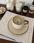 Rustic ivory mug on a saucer, placed on a fashion magazine, alongside candles and decor on a wooden table.