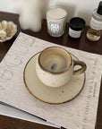 Stylish ivory mug with textured porcelain finish on a saucer, set on a wooden table beside decorative candles and a coaster.