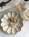 Porcelain flower-shaped jewelry dish with gold rings and pearl necklace on a marble surface alongside perfume bottles and a book.