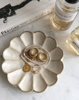 Porcelain flower-shaped jewelry dish with gold rings and pearl necklace on a marble surface alongside perfume bottles and a book.