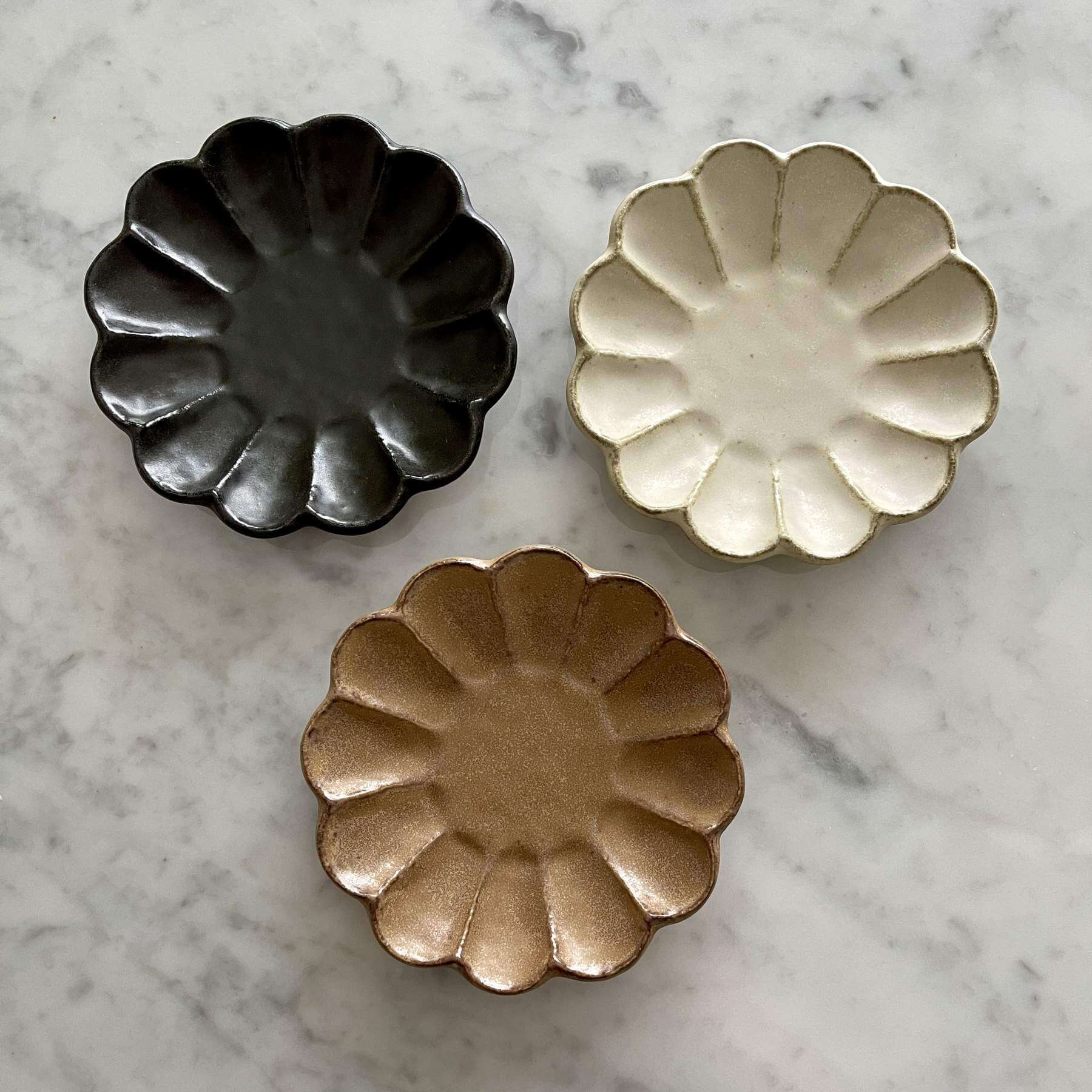 Three decorative flower-shaped porcelain plates in black, cream, and bronze colors, arranged on a marble surface.