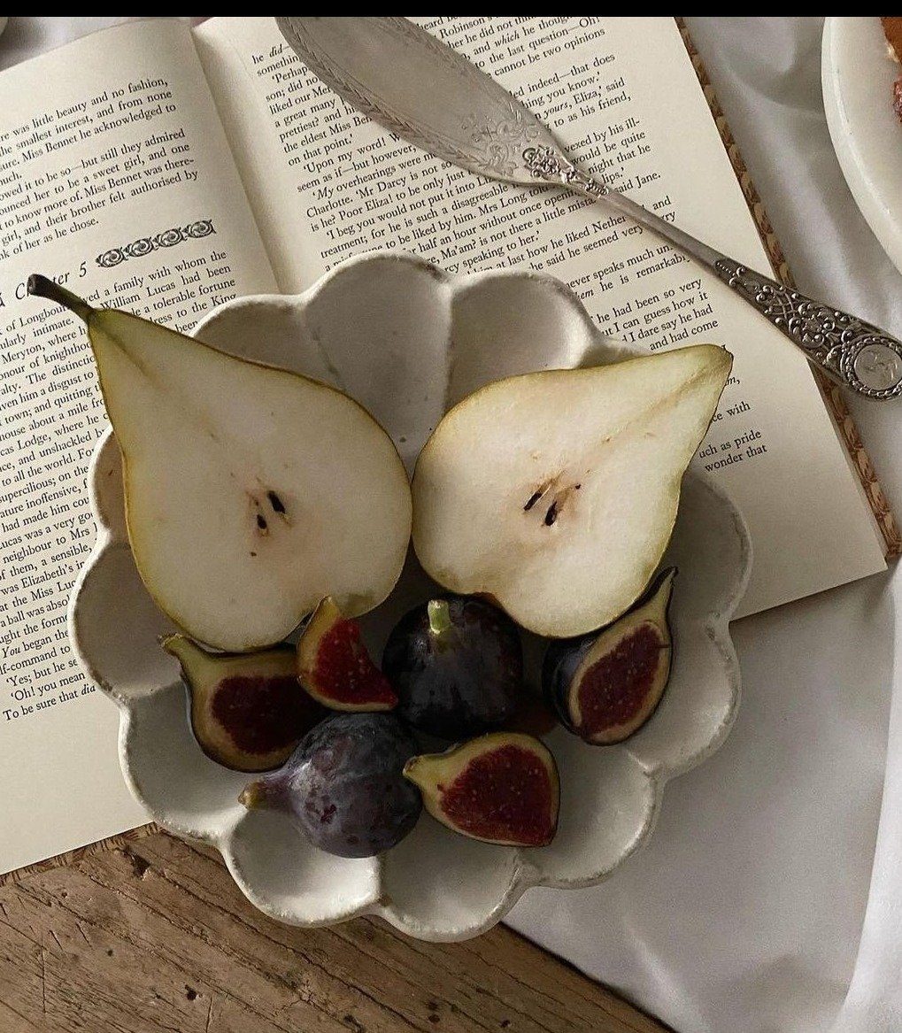 Sliced pear and figs in a scalloped ceramic bowl beside an open book with a vintage knife on a wooden surface.