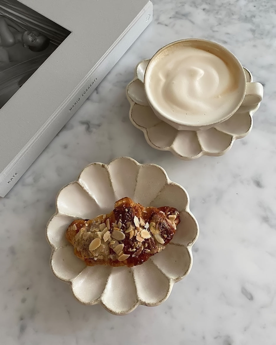 Rinka porcelain soup cup with coffee on a flower-shaped plate next to a matching plate with a pastry on a marble surface.