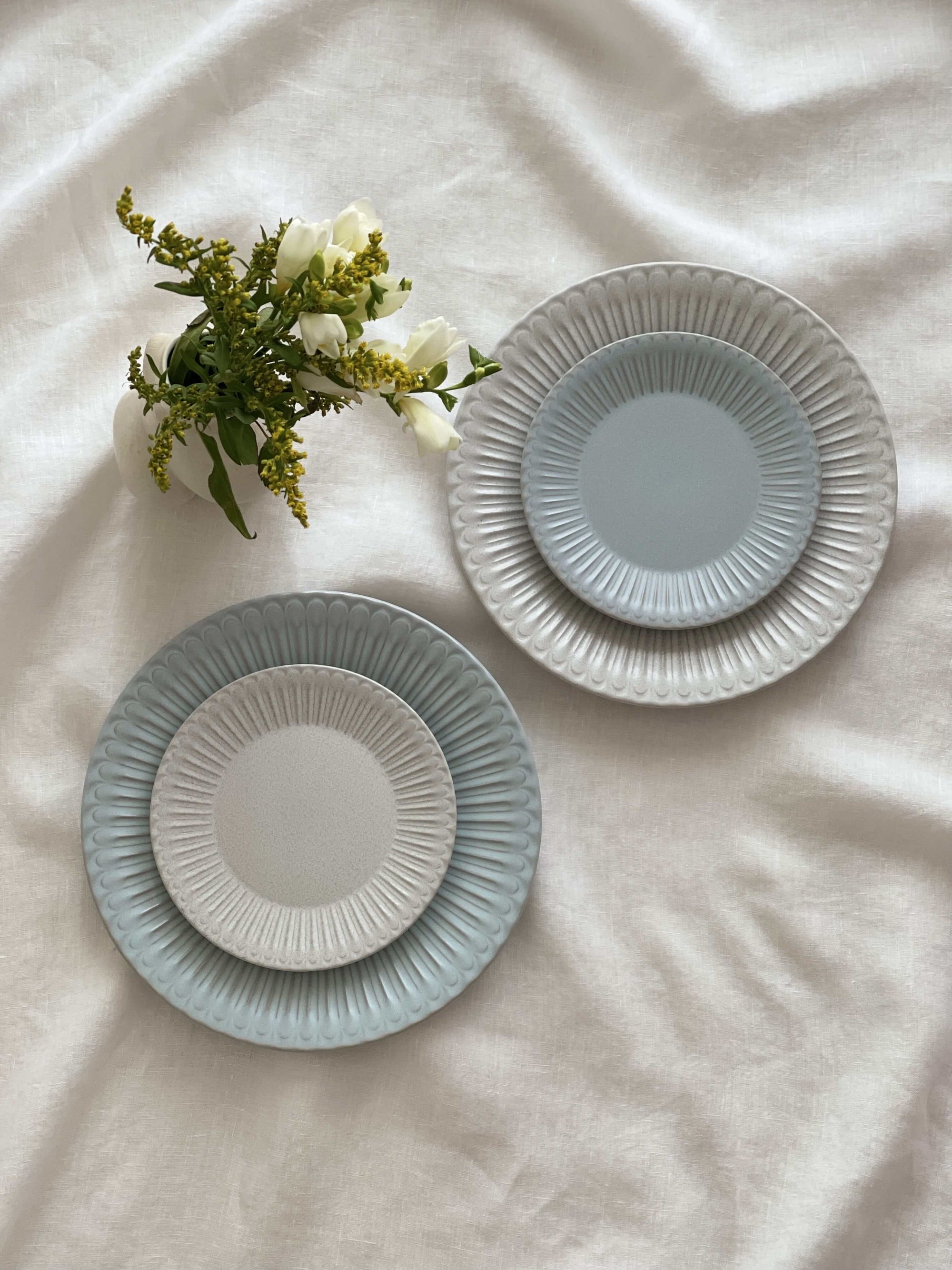 Two Storia Plates 24cm on white cloth, featuring antique-inspired lace edges and flecked lacquer finish beside a bouquet of flowers.