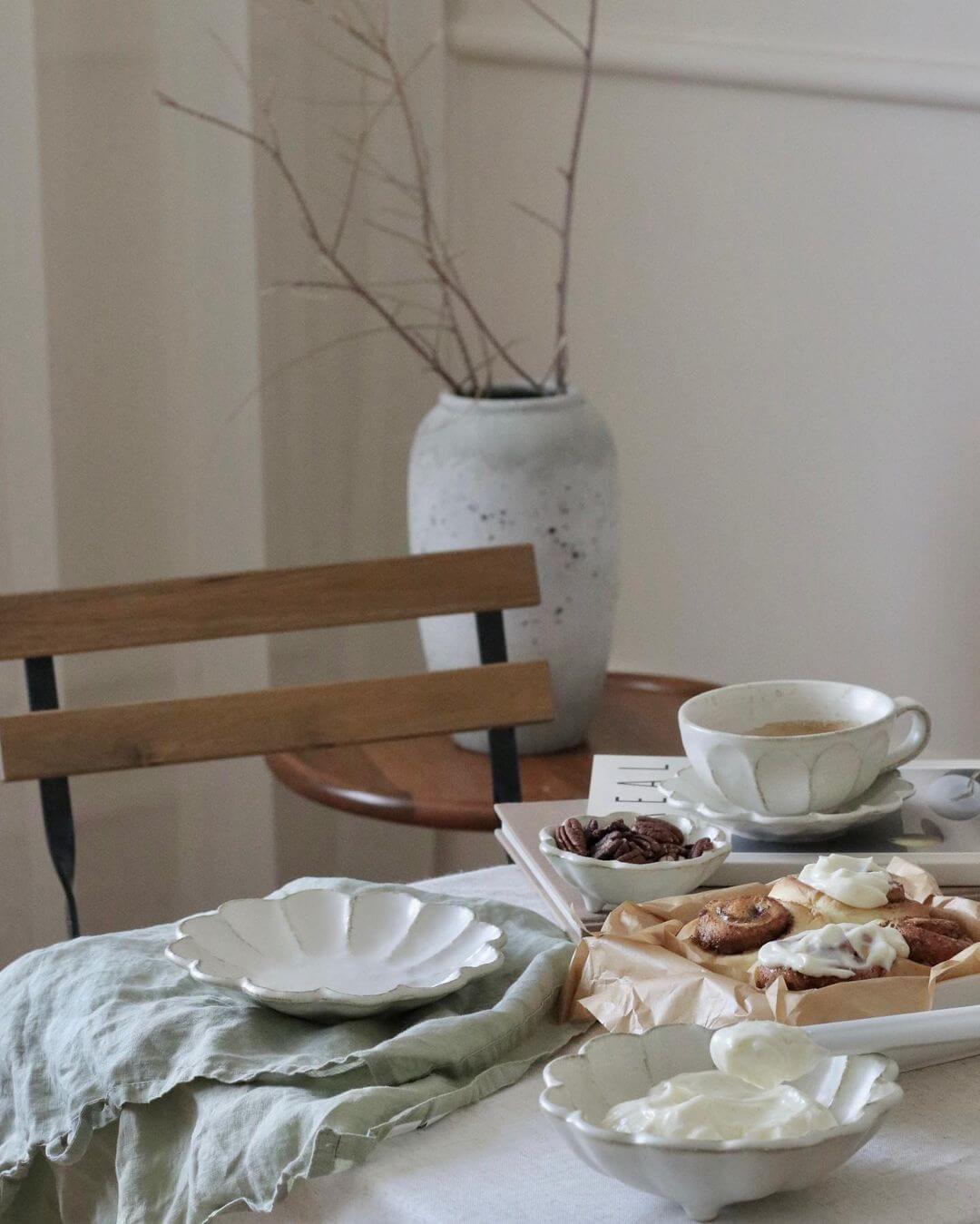 Elegant afternoon tea set with white Rinka plates and cups, featuring pastries and a rustic table setting with a vase in the background.