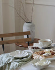 Elegant afternoon tea set with white Rinka plates and cups, featuring pastries and a rustic table setting with a vase in the background.