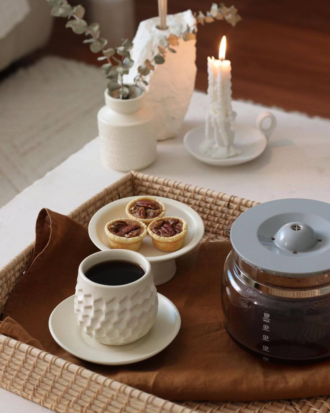 Coffee cup and pastries on a wicker tray with a white vase and candle in the background.