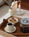 Coffee cup and pastries on a wicker tray with a white vase and candle in the background.