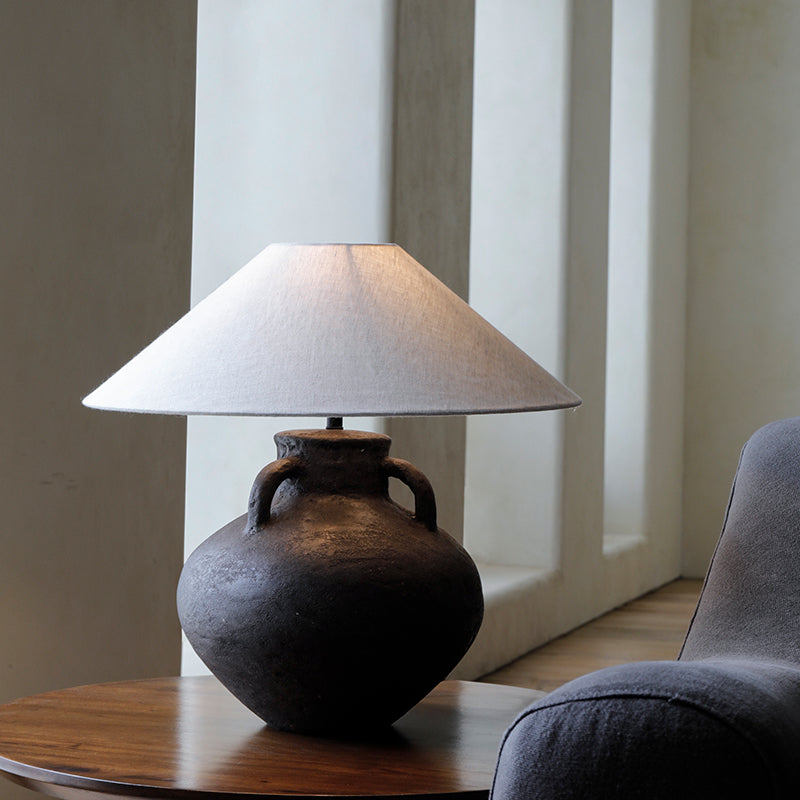 Handmade ear table lamp with pottery base and white shade, providing soft warm light on a wooden table in a cozy room.