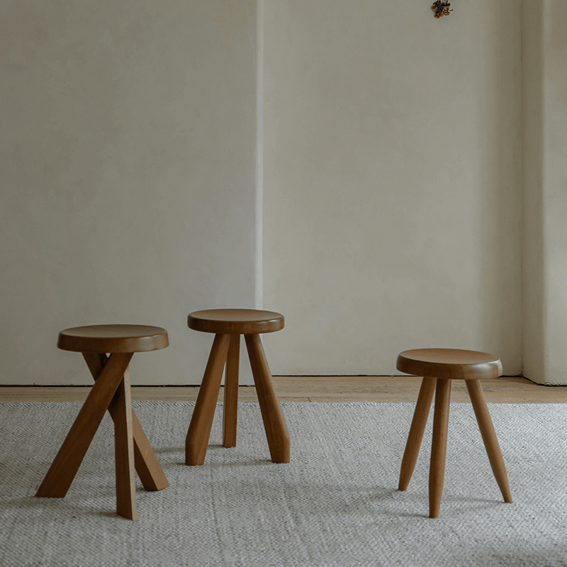 Three Meribel stools in teak wood, inspired by Alpine architecture, showcasing manual polishing and charcoal burning on a neutral rug background.
