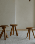 Three Meribel stools in teak wood, inspired by Alpine architecture, showcasing manual polishing and charcoal burning on a neutral rug background.