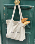 Beige tote with "LoveÉcru" logo hanging on a green door, holding two fresh baguettes.