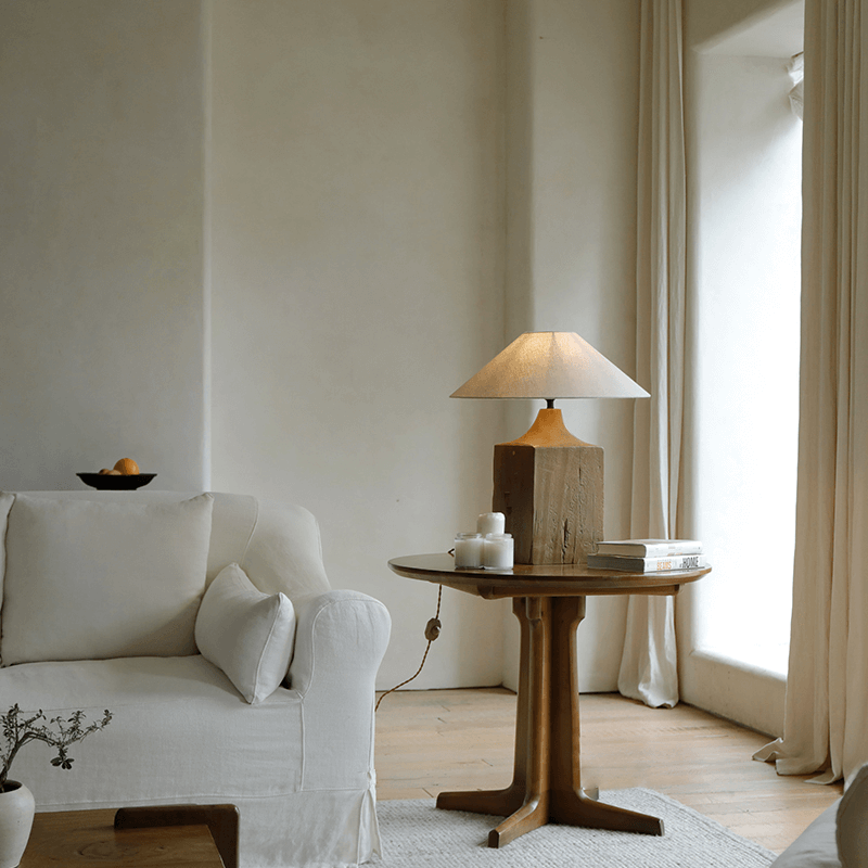 Old wood table lamp with linen shade on wooden table in cozy living room with soft beige couch and large window.